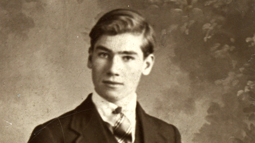 Sepia toned black and white photograph of young man with mid-dark hair wearing suit and tie in front of photographer's studio backdrop