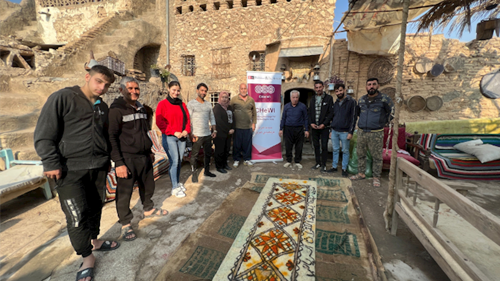 A group of workshop participants proudly displaying a wet-felted rug made during a workshop. They are standing outside a historic museum building in northern Iraq.