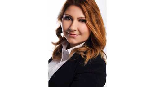 A headshot photograph of Nisreen Ameen, a light skinned woman with brown orange hair, wearing a dark suit