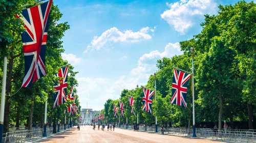 A photograph of The Mall leading towards Buckingham Palace in London. The road is lined with trees covered by green leaves with Union Jack flags hanging from evenly spaced flagpoles. about halfway down the road is a group of people. Above the sky is blue with some white clouds