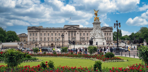 A photograph of Buckingham Palace in London with the Fountain Sculpture at the end of The Mall in front of it. Buckingham Palace is a large Victorian building, built in a Palladian style in light coloured stone.