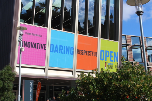 A photograph showing a close up of the Emily Wilding Davison Building on campus, Across the bottom of the windows are brightly coloured posters highlighting the  words of the new RH2030s Values - Innovative, Daring, Respectful, Open