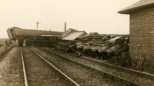 Sepia toned photograph of train derailed from tracks and lying on it's side. Men inspect derailed carriages further down the rail tracks